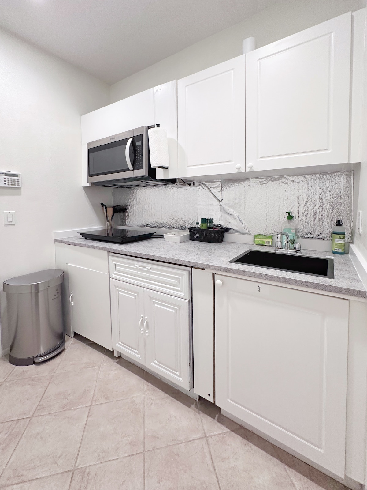 A simple kitchen is equipped with essential appliances, including a microwave and a compact stovetop. White cabinetry offers storage space, while a sink is positioned next to a countertop. A small trash bin is visible on the floor, accompanied by various kitchen supplies.