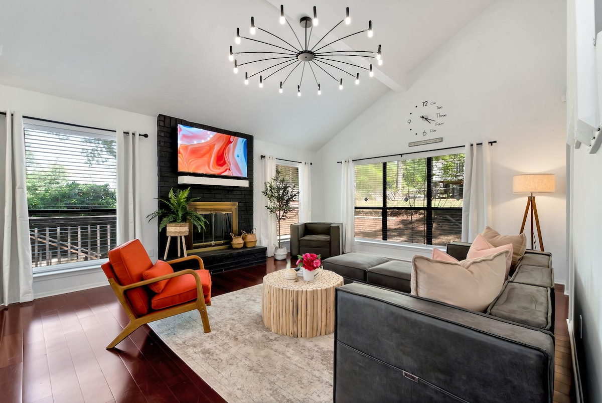 The primary living room features an open layout with large windows offering natural light. A comfortable seating arrangement includes a sectional sofa and accent chairs. A modern chandelier hangs above a central coffee table, while a fireplace and wall-mounted television provide cozy entertainment options.