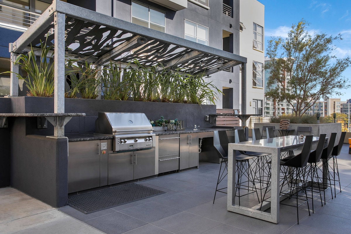 An outdoor grilling area features a built-in stainless steel barbecue surrounded by greenery. A large dining table with modern black chairs is positioned nearby, providing ample space for outdoor dining and socializing.
