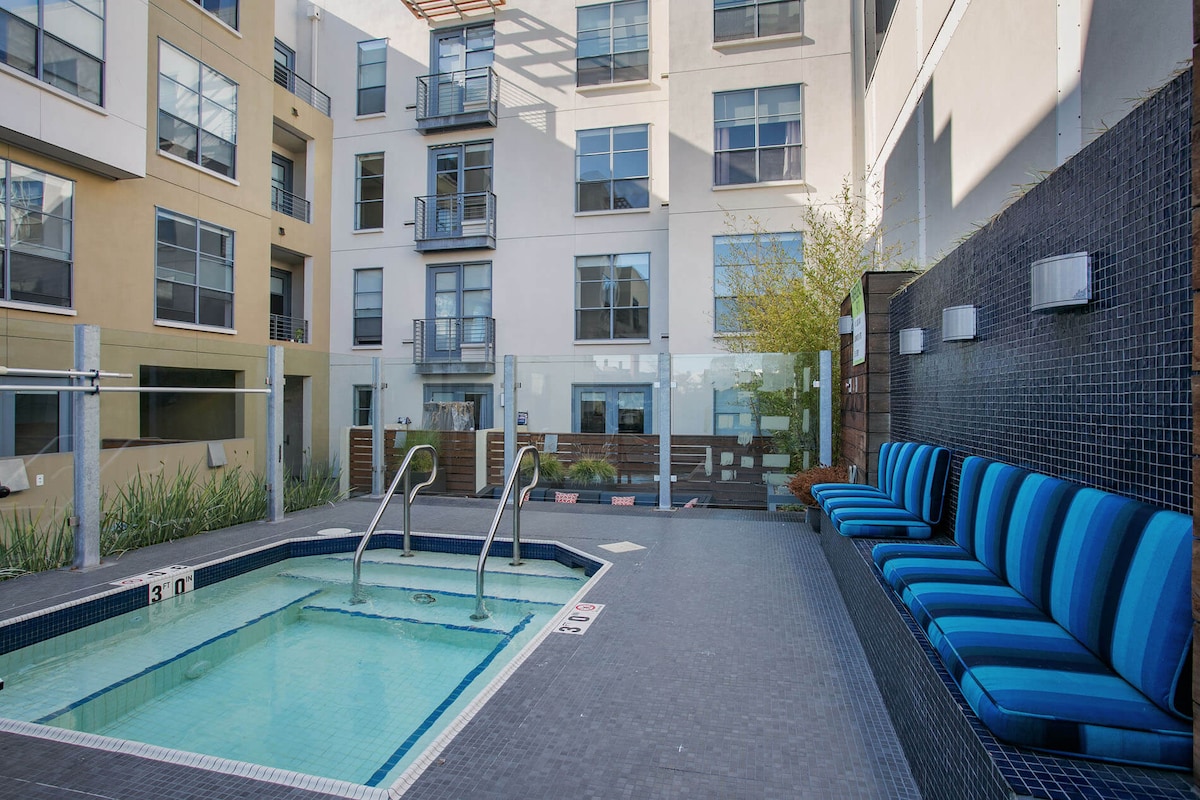 A shared outdoor pool area features a small rectangular pool with steps for entry. Surrounding the pool are modern blue and white lounge chairs. Large windows of the adjacent buildings reflect natural light and greenery, creating a relaxed and welcoming atmosphere.