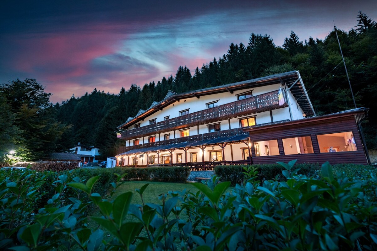 A rustic building is set against a backdrop of lush trees under a colorful twilight sky. Multiple balconies line the upper level, providing an inviting view over the well-maintained garden below. The structure's warm tones reflect the natural surroundings.