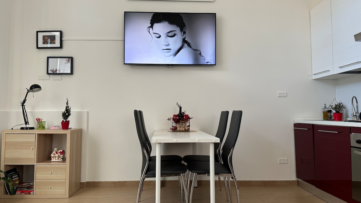 A dining area features a white table surrounded by four black chairs, creating a functional space. A mounted television is visible on the wall, with shelves displaying decorative items, adding a personal touch to the room. The kitchen area, equipped with red cabinetry, is adjacent to this space.