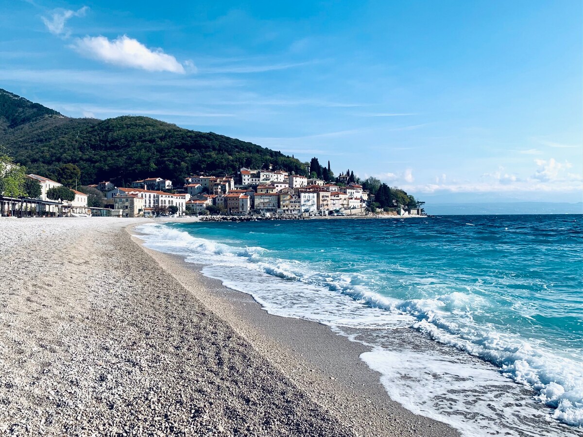 A scenic beach stretches along the coastline, with gentle waves lapping at the shore. A charming village is positioned on the hillside in the background, surrounded by lush greenery and a clear blue sky.