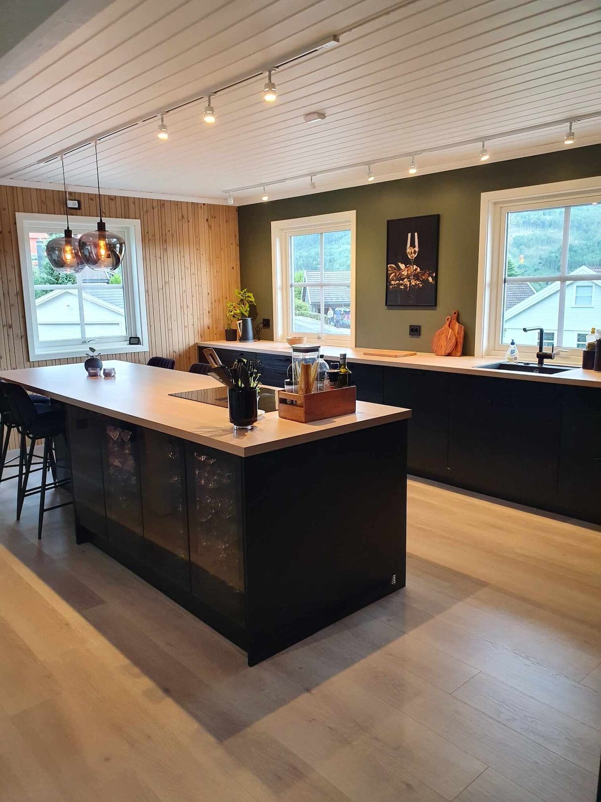 A modern kitchen features dark cabinetry with sleek countertops. Two pendant lights hang above a central island, accentuating the open space. Large windows allow natural light to fill the room, complemented by a subtle green wall and artwork enhancing the ambiance.