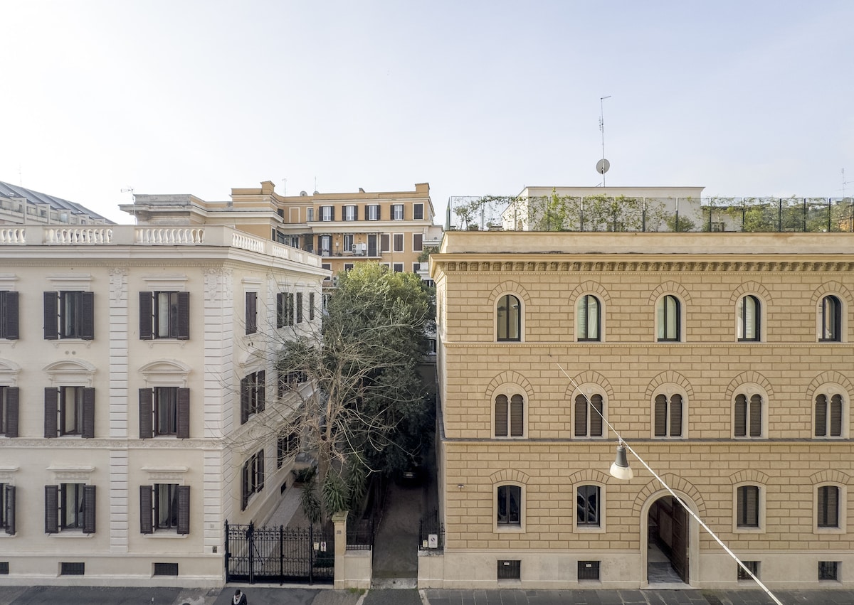 A view from the apartment showcases two adjacent buildings. The left structure features decorative elements and dark window shutters, while the right building has a textured facade and minimalistic design. Lush greenery is visible between the buildings, adding a touch of nature to the urban environment.