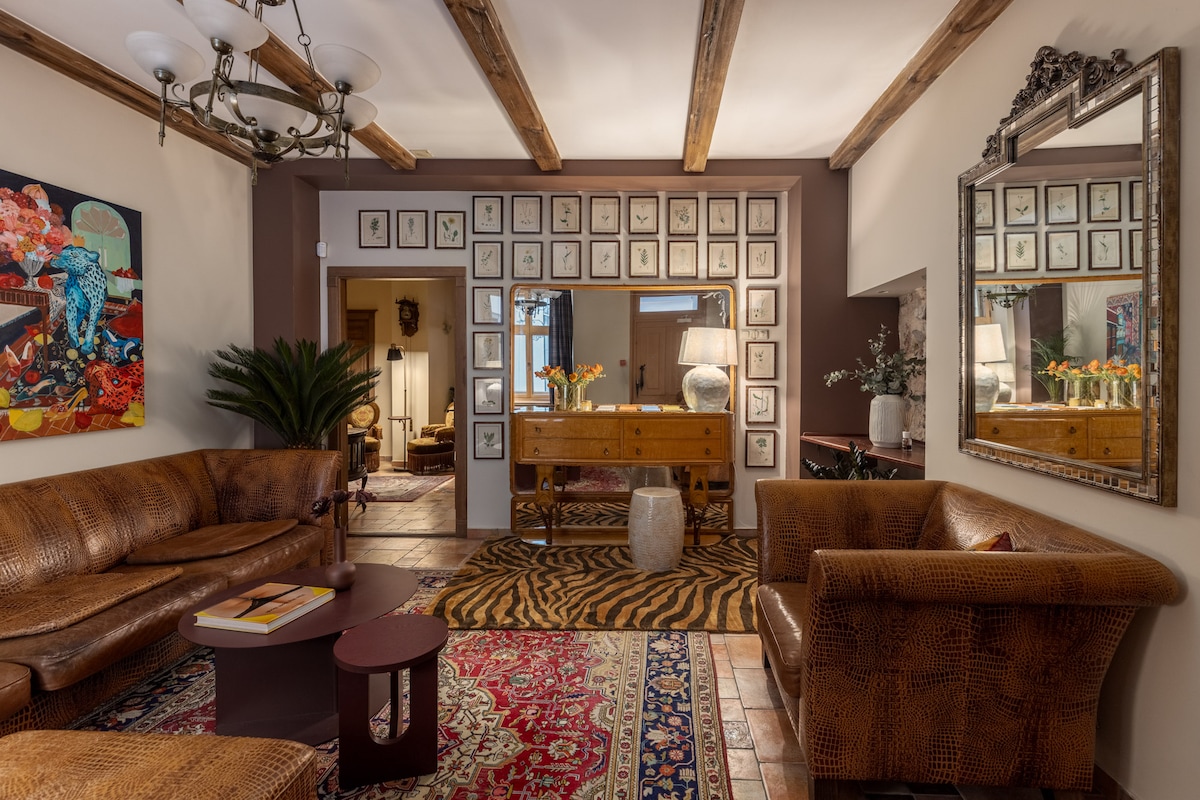 A spacious living area is highlighted by exposed wooden beams and warm-toned decor. Comfortable seating arrangements consist of leather sofas and armchairs, accompanied by a coffee table. An arrangement of framed artworks adorns the walls, reflecting a blend of contemporary and classic styles.