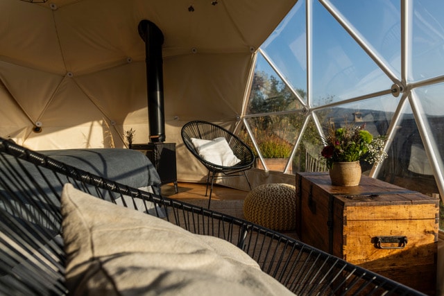 A cozy interior of a geodesic dome is highlighted, featuring a comfortable seating area with a black woven chair and a soft cushion. A vintage wooden crate serves as a table, adorned with a plant and flowers, while a stove adds warmth alongside expansive windows showcasing the outside view.