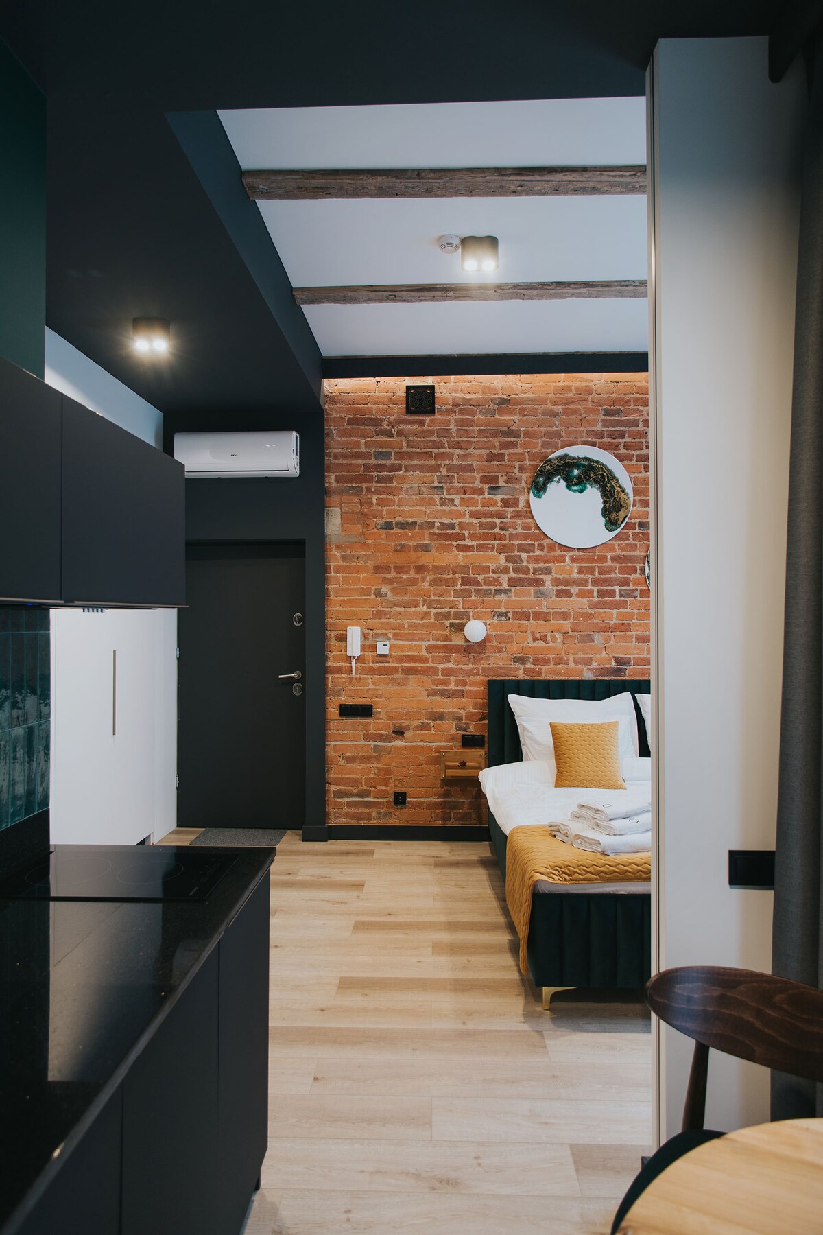 A studio space is highlighted by exposed brick walls and wooden beams. A cozy bed is dressed in neutral linens, featuring an accent pillow. A kitchenette with black cabinetry and modern appliances is visible, along with a round mirror and a small dining area.