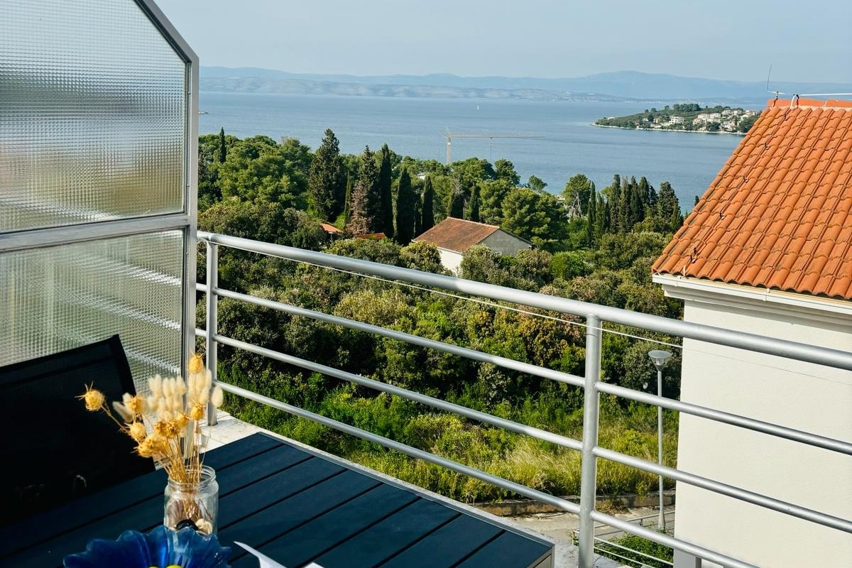 A spacious terrace is presented with a dark table adorned with a vase of dried flowers. Beyond, a panoramic view of the sea and lush greenery is visible, while a distant coastal area adds to the natural scenery.