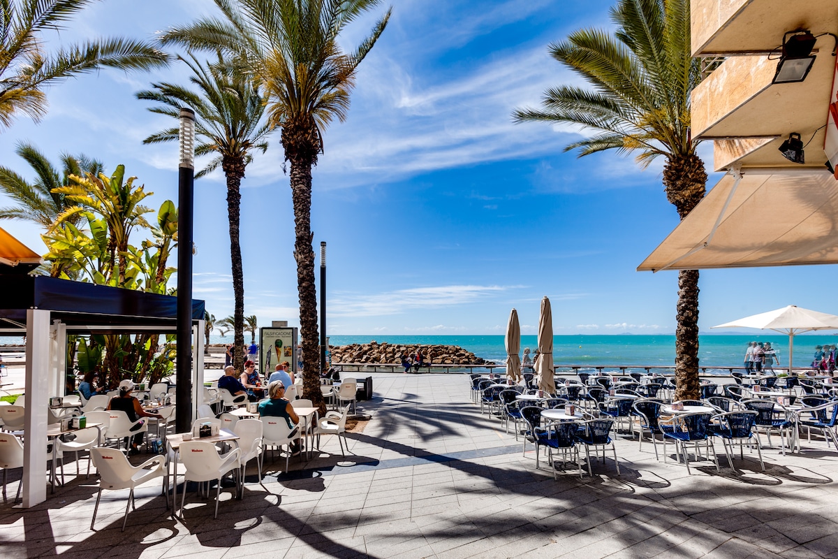 An inviting seaside promenade features numerous outdoor seating options under the shade of tall palm trees. Sunlight casts playful shadows on the paving, creating a warm atmosphere. The clear blue sky and calm turquoise waters enhance the tranquil setting, ideal for leisurely gatherings.