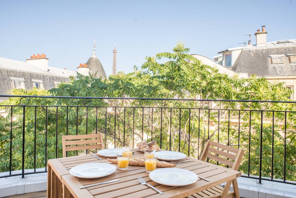 A furnished balcony showcases a wooden table set for breakfast, featuring plates, glasses of orange juice, and a basket of pastries. Lush greenery surrounds the space, while a glimpse of the Eiffel Tower is visible in the background, enhancing the serene setting.