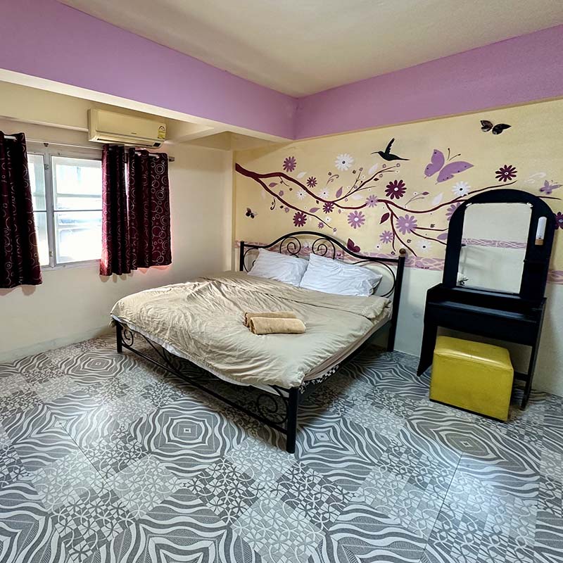 A spacious sleeping area is depicted with a large bed dressed in neutral linens. A simple vanity table with a yellow stool is positioned beside the bed. Dark curtains frame the bright window, and decorative wall art features floral designs and butterflies.