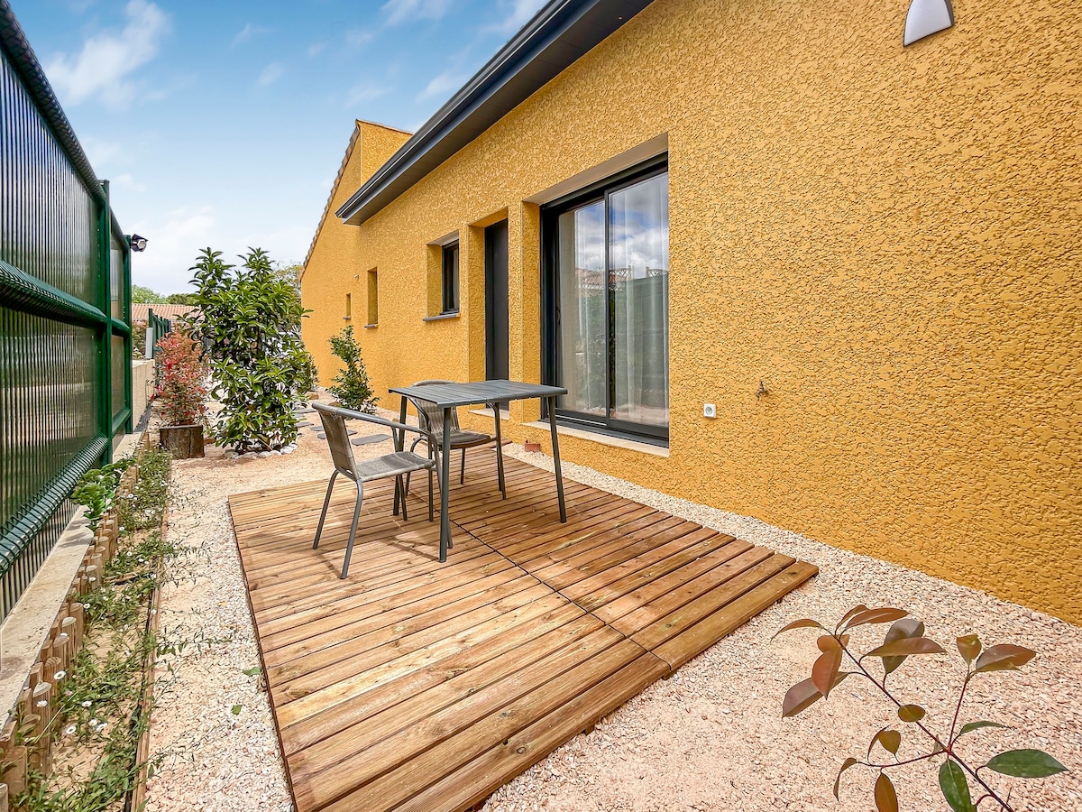 A private terrace is presented with wooden decking and a small table surrounded by two chairs. Lush greenery provides a natural backdrop, and a textured yellow wall of the building adds warmth to the setting.