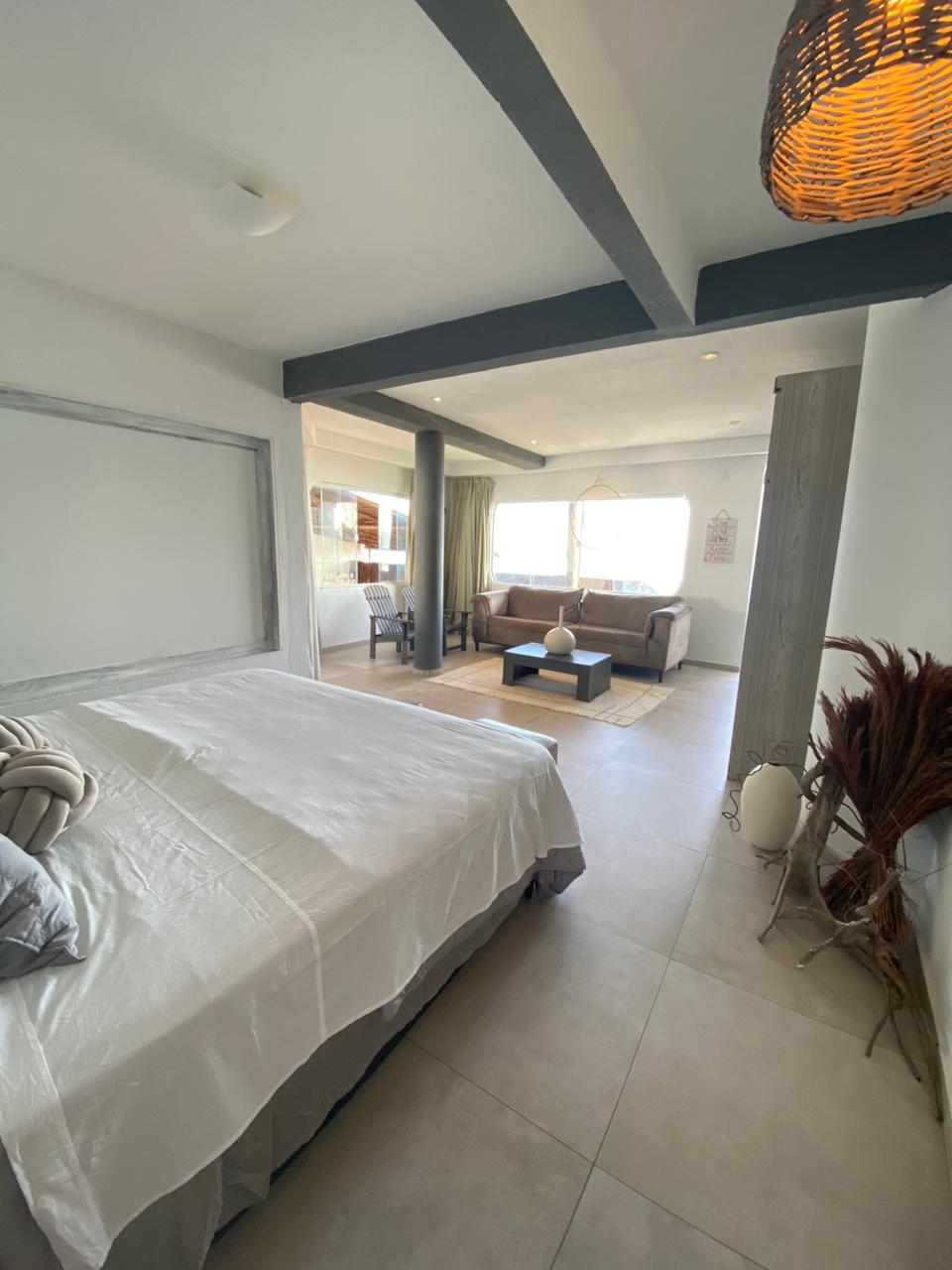 A cozy room is featured, featuring a spacious bed draped with light bedding. A comfortable seating area with a couch and chair is visible, adjacent to a large window allowing natural light. Neutral tones create a calming ambiance throughout the space.