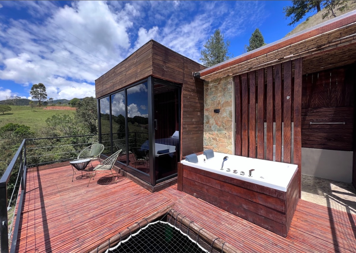 A private terrace showcases a jacuzzi tub and two stylish chairs, surrounded by wooden decking. Large glass doors provide views of the lush landscape, while natural stone accents add texture to the cabin's exterior.