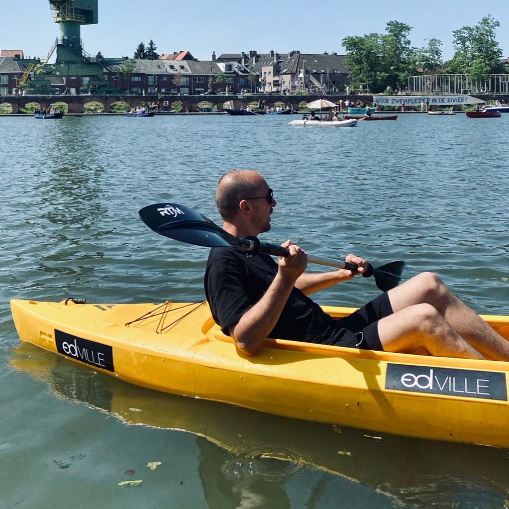 A bright yellow kayak is seen afloat on calm waters, with a person paddling. The backdrop features historic buildings and greenery along the shoreline, while a distant structure, appearing as a watchtower, adds interest to the scene.