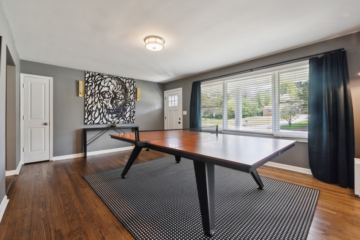 A game room is featured with a sleek pingpong table set on a patterned rug. Natural light filters through large windows adorned with room darkening shades, enhancing the modern decor highlighted by an abstract wall art piece.