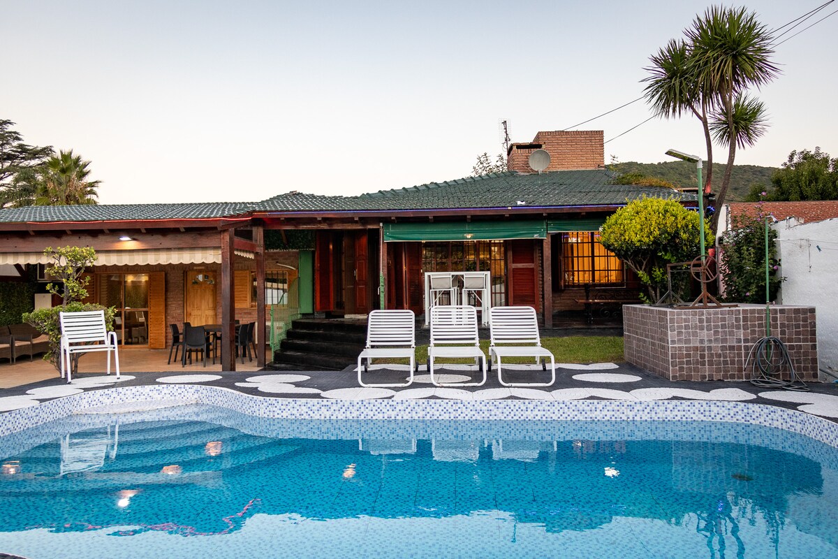 A refreshing pool reflects the evening light outside a charming house featuring an open patio area. White lounge chairs are positioned around the pool, while green foliage and smooth stone paths enhance the inviting space.