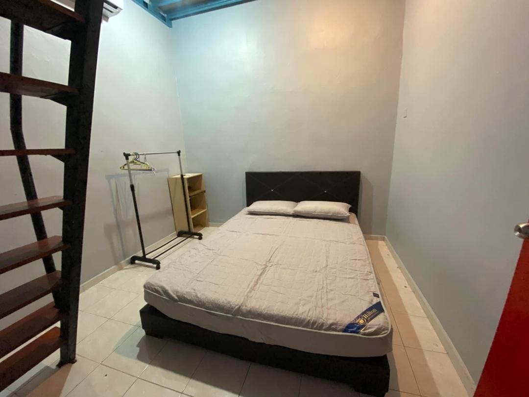 A simple bedroom is showcased, featuring a spacious bed with neutral bedding against a light grey wall. A clothing rack and a small shelf are positioned nearby, while a staircase leading to another level can be seen on the left.
