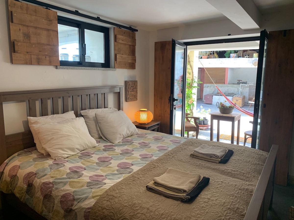 A serene bedroom presents a spacious bed, adorned with a colorful patterned quilt and neatly arranged towels. Natural light enters through sliding glass doors, leading to an outdoor area with greenery. Wooden accents are featured in the decor, and soft lighting from a bedside lamp enhances the ambiance.
