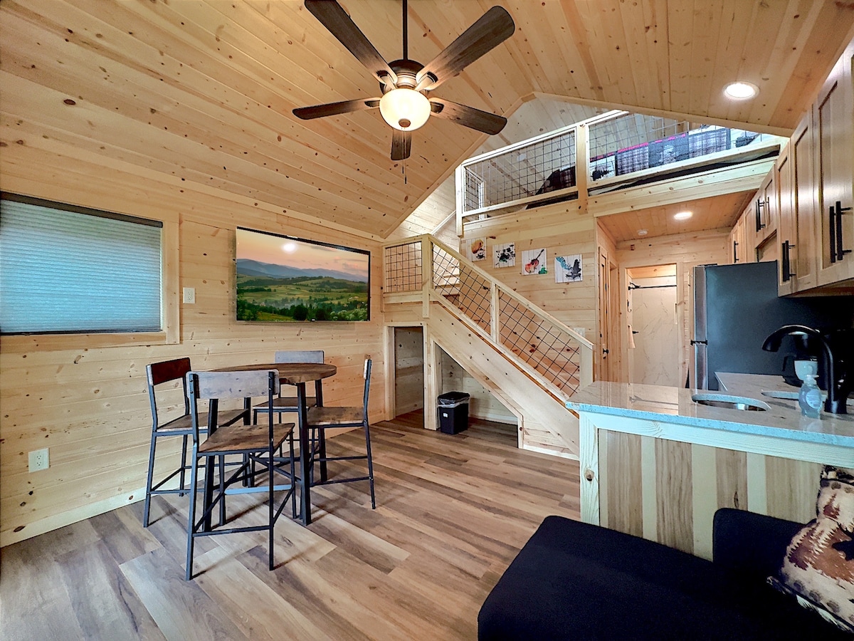 An open living space features a wooden ceiling and walls, complemented by a ceiling fan. A dining table with four chairs is situated near the kitchen area, while a cozy sofa is positioned in one corner. A staircase leads to a lofted sleeping area above.