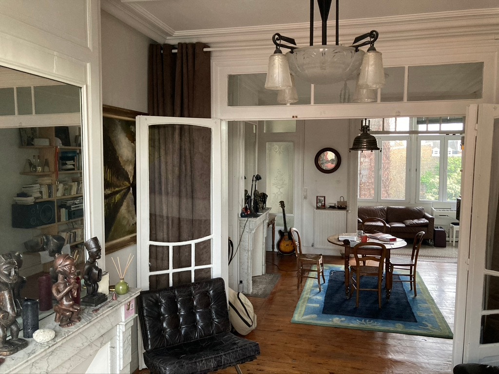 A view of the living area reveals a blend of modern and vintage elements, featuring a round dining table surrounded by wooden chairs. Light enters through large windows, while a cozy seating area with a low chair and decorative sculptures enhances the space's charm.