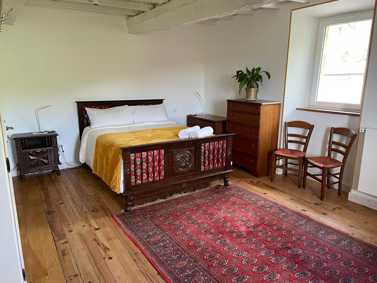 The room features a spacious bed adorned with an elegant yellow coverlet. A colorful wooden headboard adds a touch of charm, complemented by a traditional area rug. A wooden dresser and two chairs are arranged thoughtfully, with a plant positioned on the dresser beside a window.