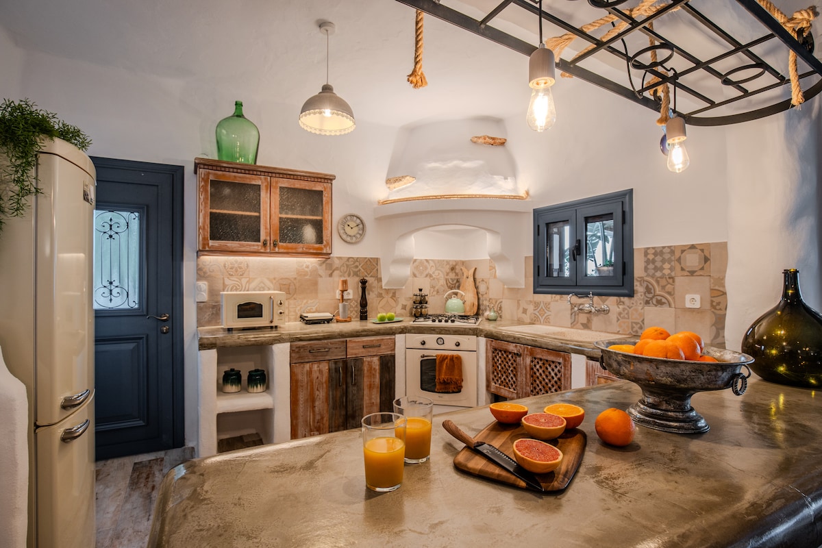 A rustic kitchen features wooden cabinetry and ceramic tiles. A vintage refrigerator and modern appliances are included. A large countertop holds cut oranges and a bowl, while glasses of juice sit nearby. Warm lighting from hanging fixtures enhances the inviting atmosphere.
