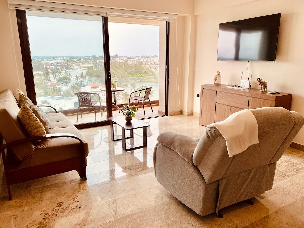 A comfortable living area is presented, featuring a spacious sofa and a recliner. A glass coffee table sits in the center, with a small plant adding a touch of greenery. Large windows provide views of the surrounding landscape, and a flat-screen television is mounted on the wall.