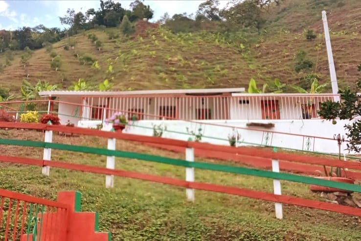 A hillside home is surrounded by lush greenery and a vibrant landscape. A white exterior is complemented by a decorative railing painted in red and green. Potted flowers add color to the entrance, while the mountainous terrain creates a natural backdrop.