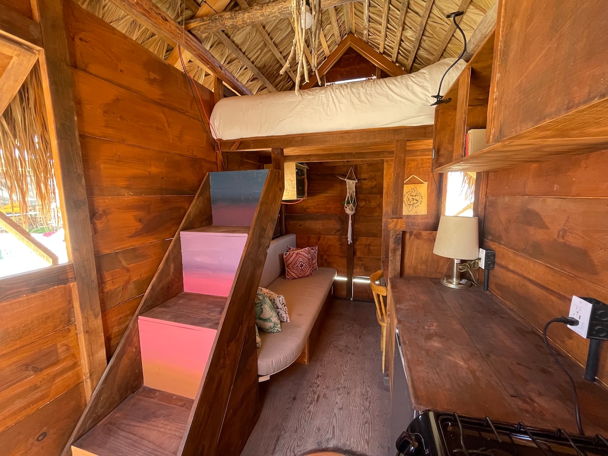 The interior features warm wooden walls and a cozy lofted sleeping area accessed by colorful stairs. A comfortable sofa sits below, complemented by a small workspace with a lamp and kitchen area equipped with essential appliances and utensils.