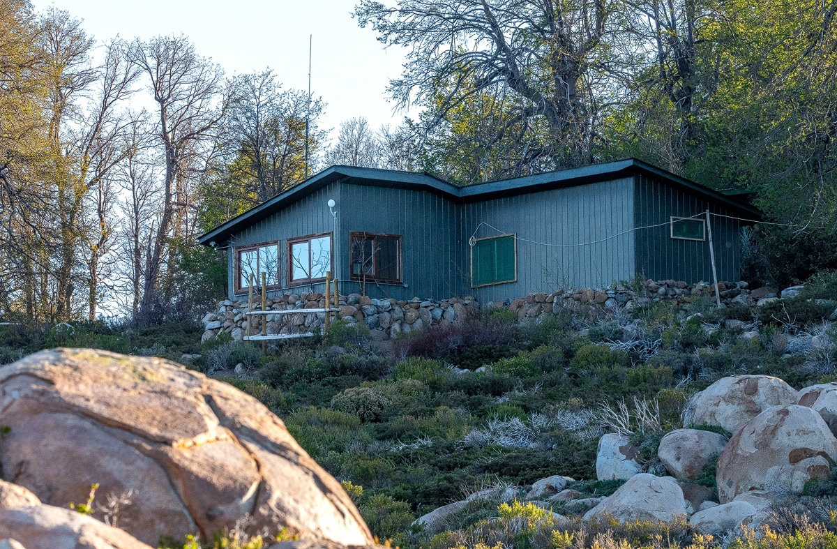 A unique cabin is nestled among rocky terrain and greenery. The structure features large windows and a stone foundation, blending naturally with its surroundings. Sunlight highlights the textures of the walls and the landscape, creating a connection with nature.