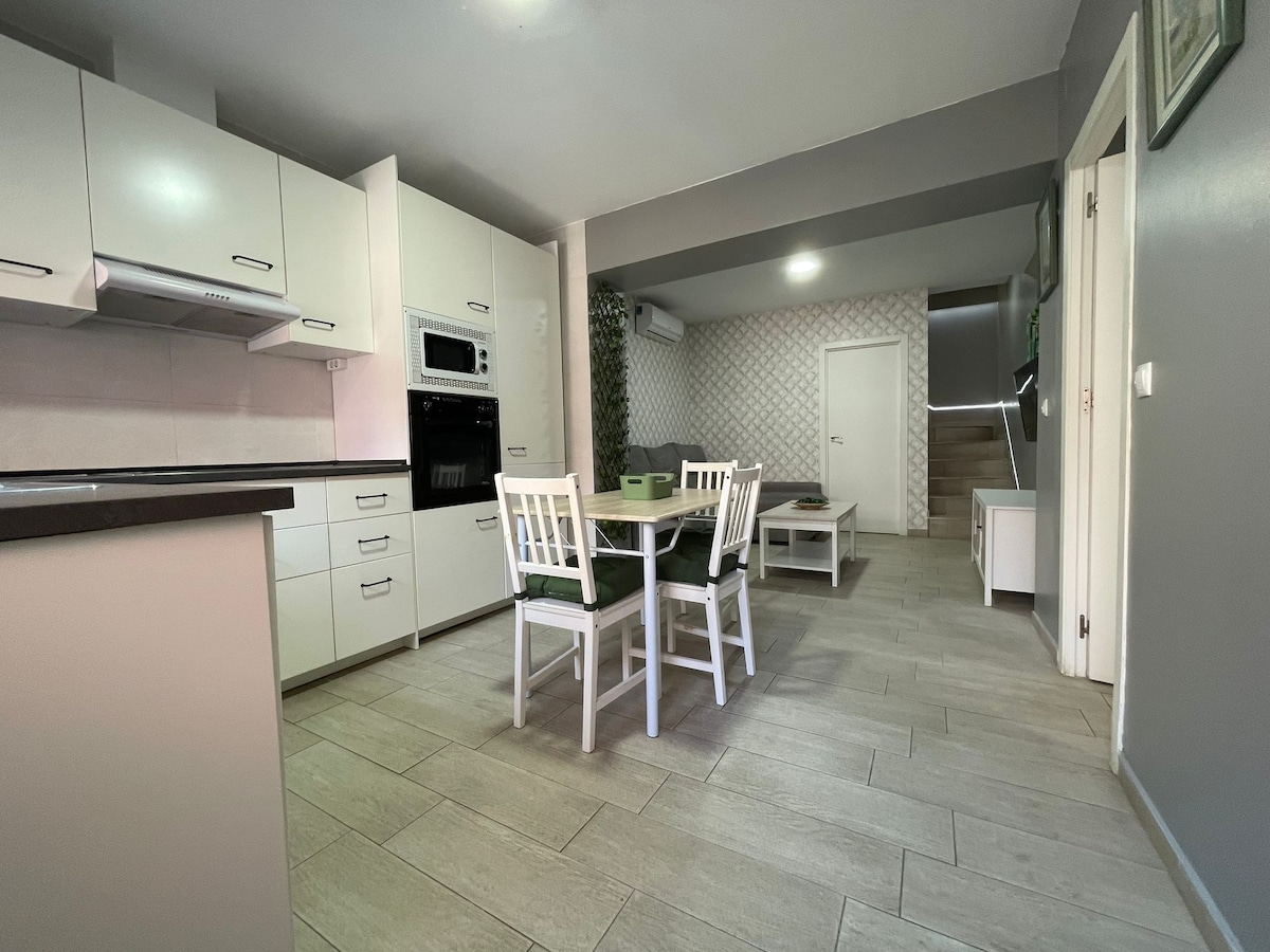 A bright kitchen area features white cabinetry and modern appliances. A dining table with four chairs is positioned centrally, accompanied by a small seating area. Neutral tile flooring extends throughout, creating a spacious, open-concept layout that connects to the staircase and living spaces.