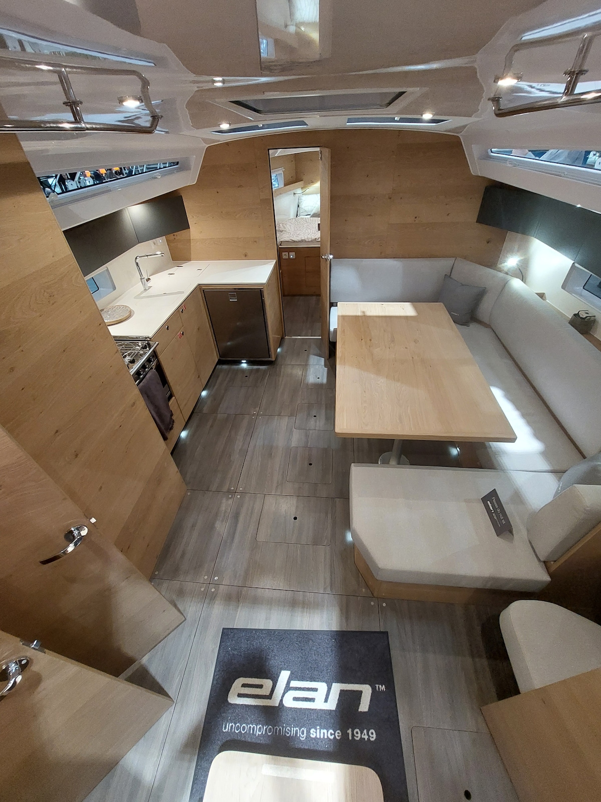 The interior space of the boat showcases a modern design with natural wood accents. A kitchen area is visible with essential appliances. A dining table surrounded by comfortable seating is positioned centrally, complemented by light-colored cushions and soft lighting, contributing to a cozy atmosphere.