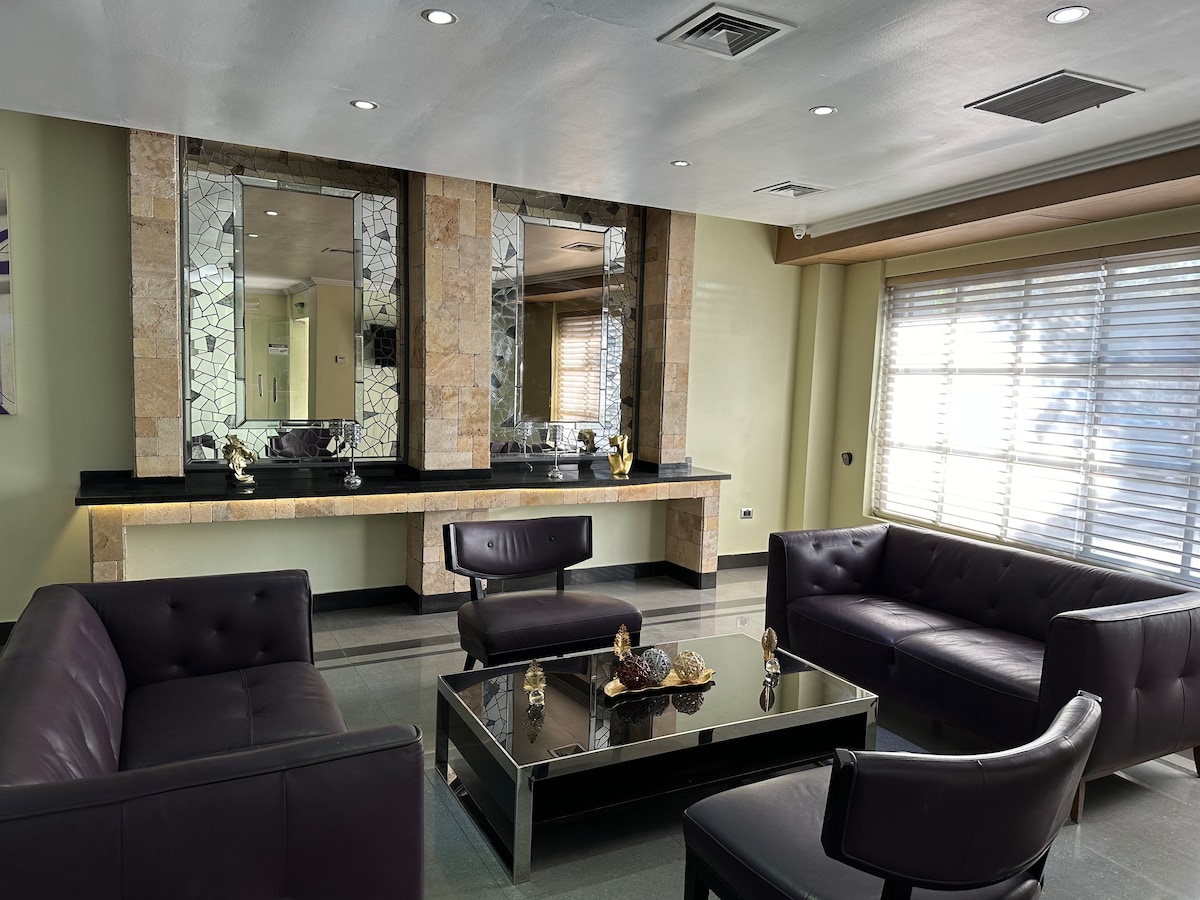 A modern lounge area features sleek black sofas arranged around a glass coffee table. Light reflects off the mirrored wall, enhancing the spacious feel. A large window with horizontal blinds provides natural light, complementing the contemporary design.