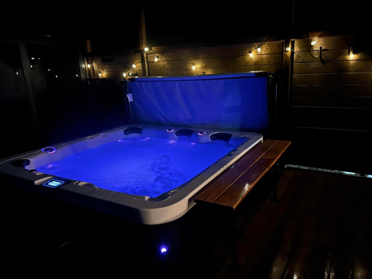 A private hot tub is illuminated with soft blue lighting, creating a serene atmosphere. The surrounding area features wooden paneling and warm string lights, enhancing the inviting ambiance for relaxation. A wooden bench is positioned nearby, offering a space for comfort.