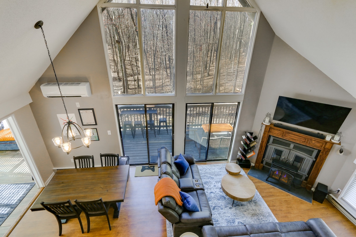 An aerial view showcases an open living space with large windows allowing natural light to flood in. A dining table with dark chairs sits near the windows, while a comfortable seating area features leather recliners and a modern coffee table, all surrounded by a serene wooded backdrop.