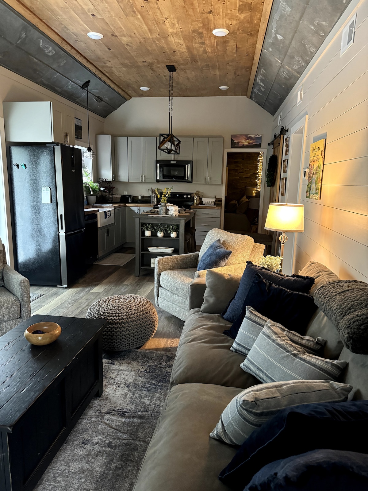 A comfortable living space is highlighted by a plush sofa adorned with decorative pillows. A stylish coffee table sits at the center, accompanied by a cozy pouf. The kitchen area features modern appliances, while soft lighting creates a relaxing ambiance.