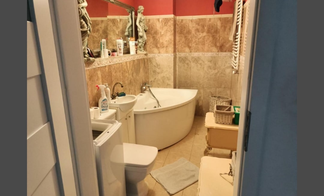 A bathroom is showcased, featuring a curved bathtub and a separate toilet. A sink is positioned adjacent to the bathtub, with various toiletries arranged on the countertop. Decorative elements include a neutral tile design and wall accents, creating a functional and aesthetically pleasing space.