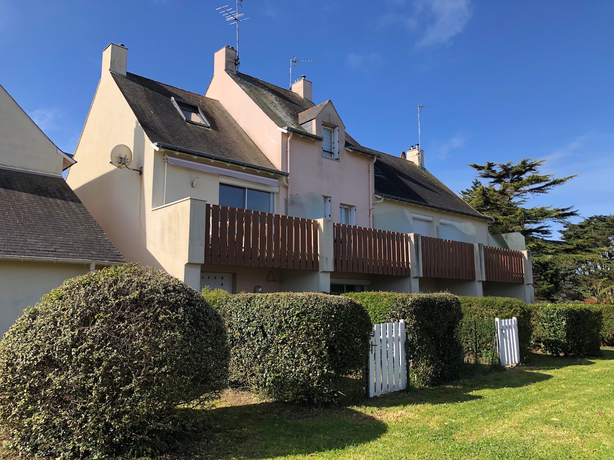 The exterior of the cottage features a two-story structure with light-colored walls and a balcony that includes wooden railings. Neat hedges surround the property, and clear skies provide a bright backdrop. The overall setting suggests a quiet and serene environment.