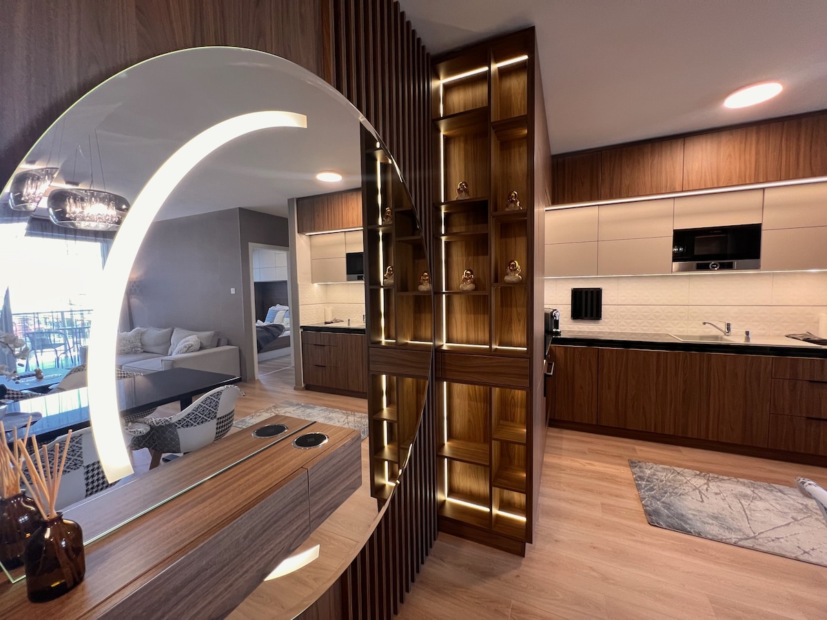 An open kitchen space features sleek cabinetry and modern appliances, illuminated by warm lighting. A stylish circular mirror adds a contemporary touch, while the adjacent living area is partially visible, showcasing cozy seating and a smooth, neutral palette.