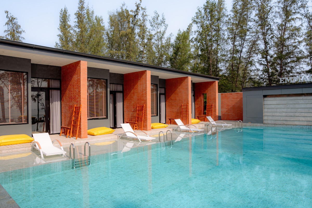 A spacious outdoor swimming pool is framed by modern flat-roofed buildings featuring large windows. Several loungers are positioned beside the pool, cushioned with bright yellow pads. Lush trees provide a natural backdrop, enhancing the serene outdoor setting.