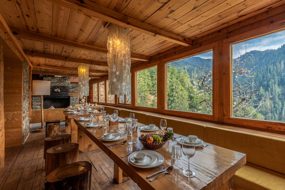 A spacious dining area showcases a long wooden table set for a meal, surrounded by large windows offering views of the mountains and trees. Natural light is filtered through multiple hanging light fixtures, enhancing the room's atmosphere.