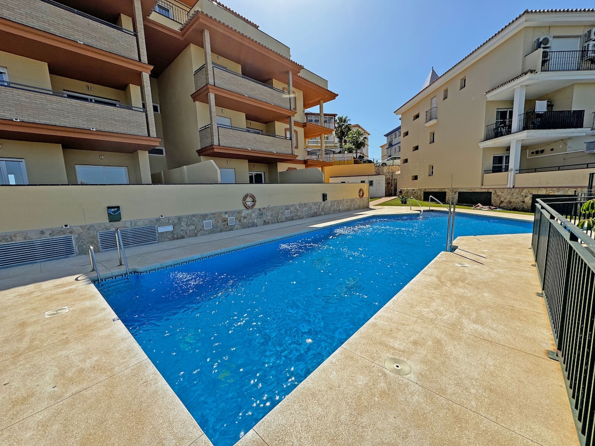 A tranquil swimming pool is surrounded by a spacious patio, featuring lounge areas and shaded sections. Sunlight reflects off the clear blue water. The pool area is bordered by low fencing, with buildings visible in the background, providing a serene atmosphere for relaxation.