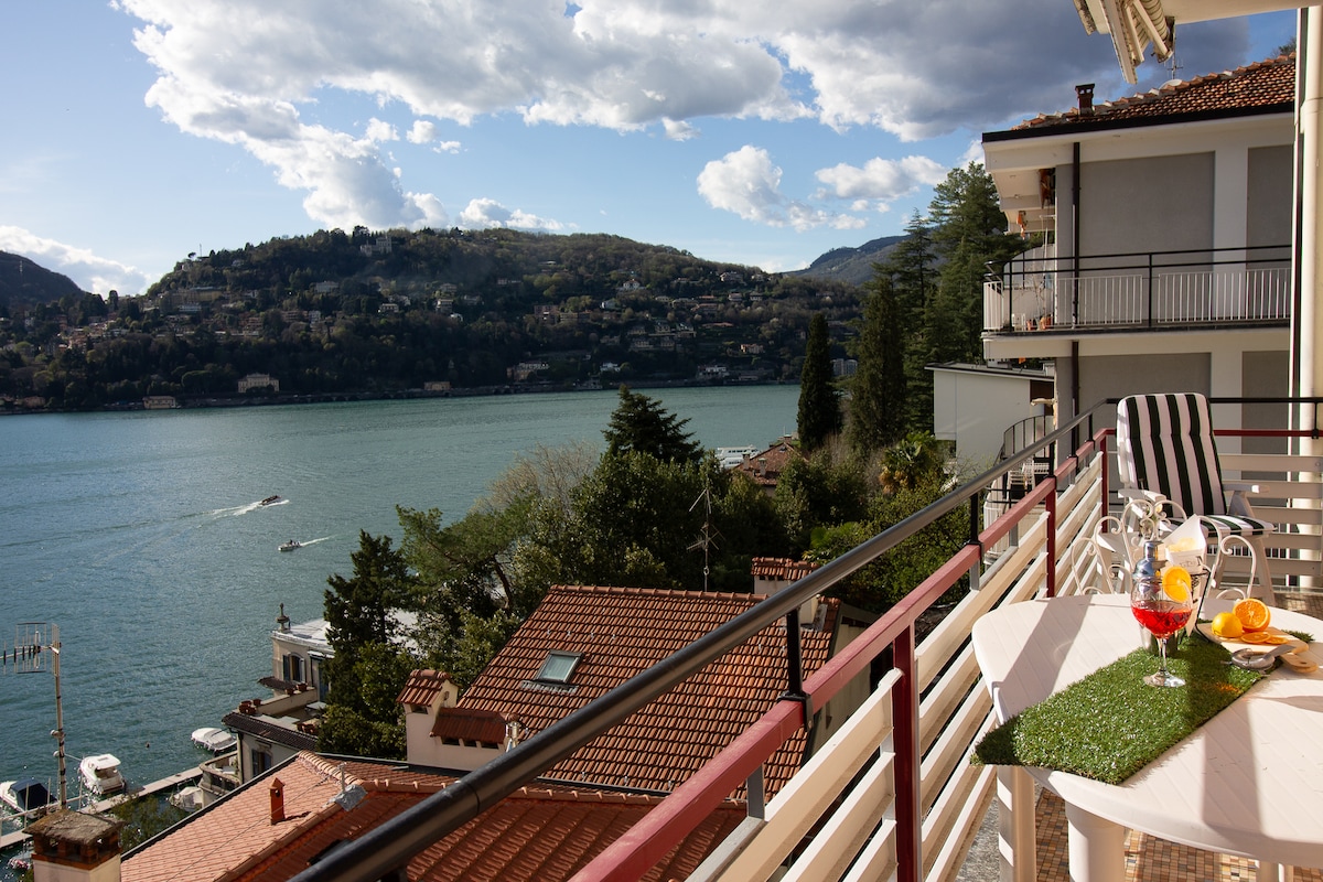 A spacious balcony is featured, overlooking a serene lake and lush mountains. A small table is set with drinks and fruits, inviting relaxation. Surrounding rooftops and greenery enhance the picturesque view, with blue skies and scattered clouds creating a tranquil atmosphere.