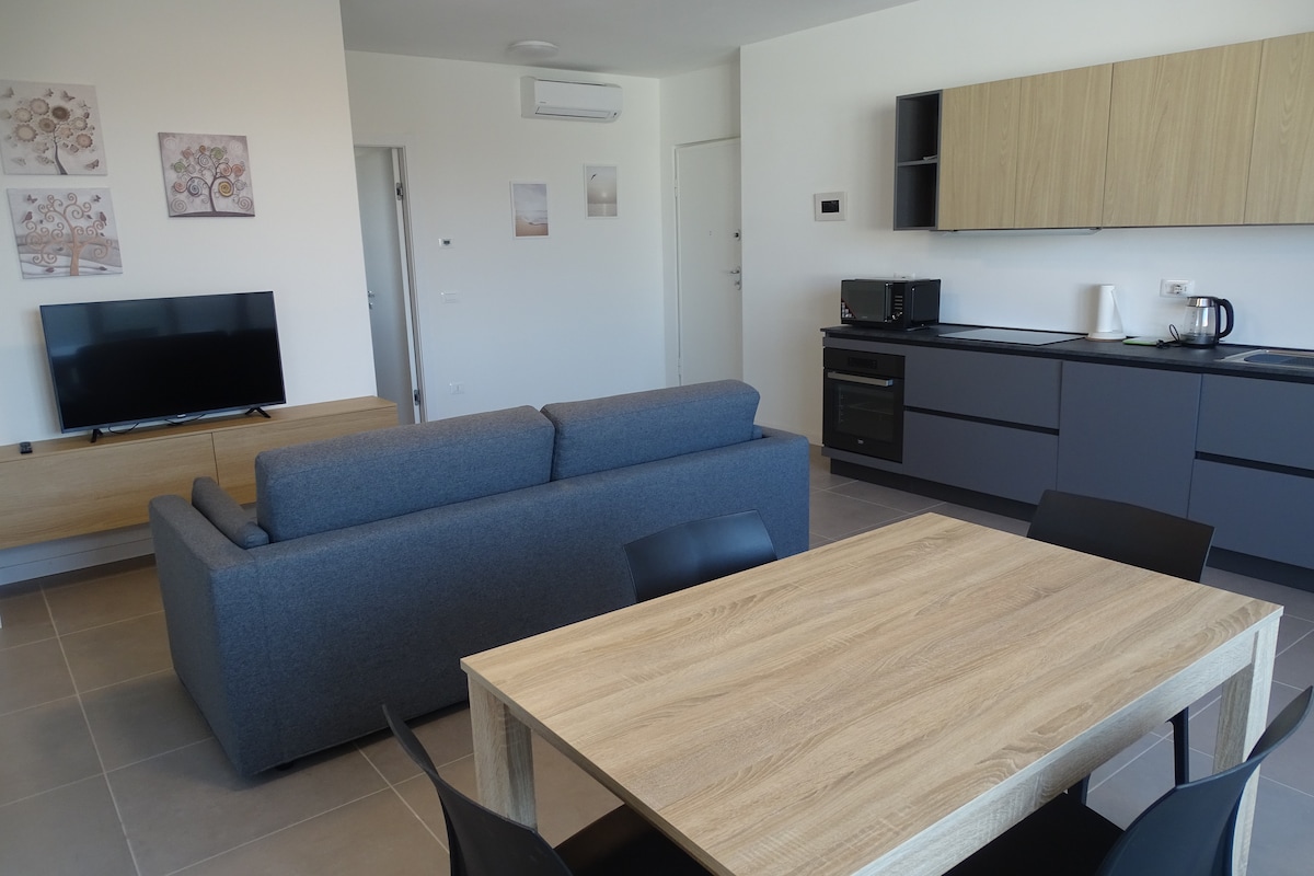 An open-plan living area features a gray sofa facing a flat-screen television. A wooden dining table accommodates several chairs, while a modern kitchen showcases integrated appliances and wooden cabinetry. Natural light enters through large windows, creating a bright and airy environment.