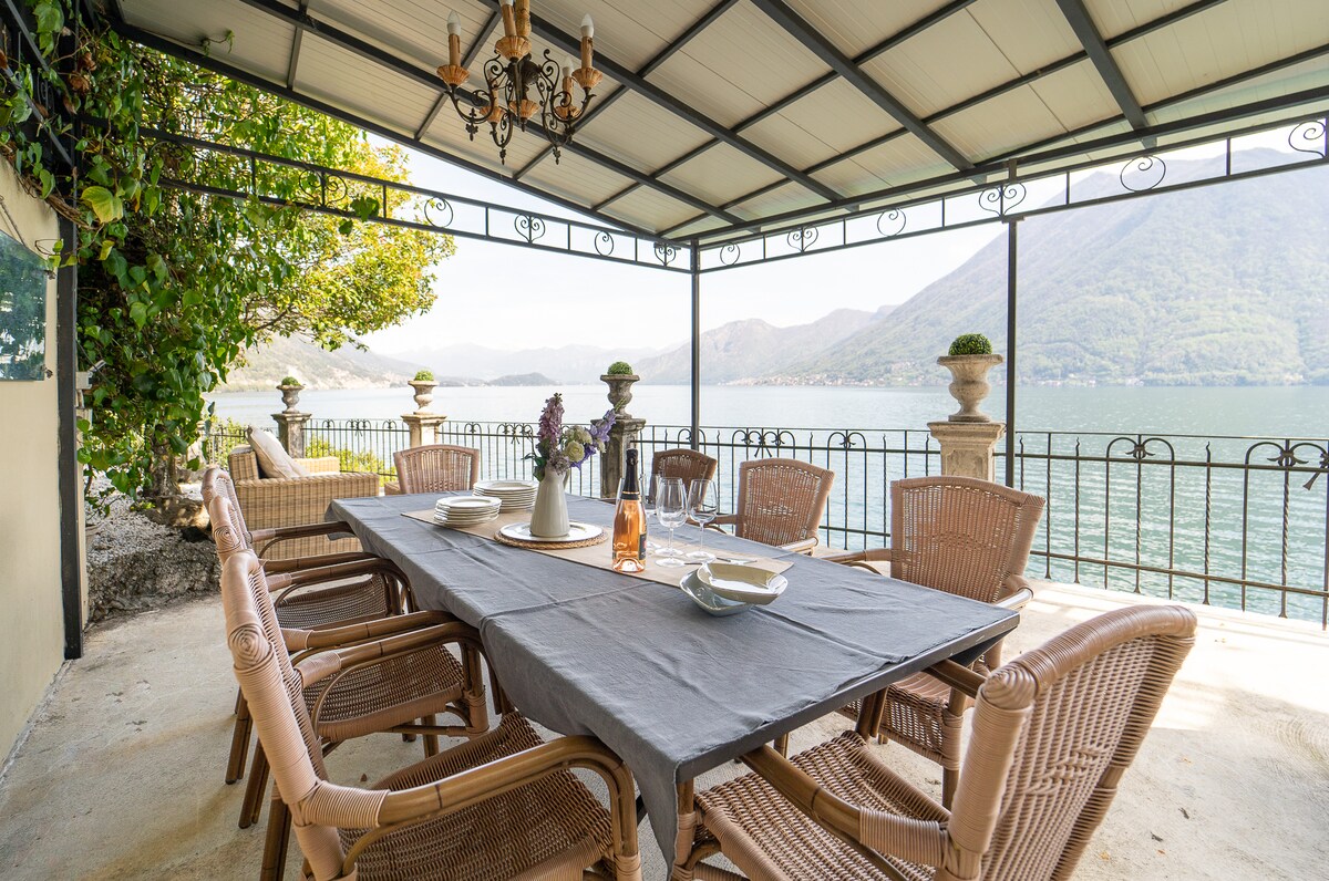 A dining area features a large table surrounded by elegant wicker chairs, set under a covered terrace. Views of the lake and mountains are visible through the railing. Soft greenery is present in the background, adding a touch of nature to the space.
