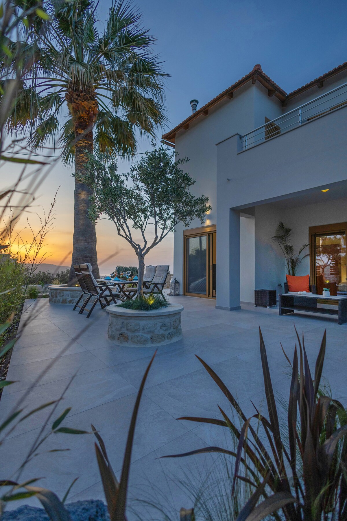 An outdoor seating area is situated beneath a palm tree, featuring a circular stone bench and a set of wooden chairs. The evening sky displays warm hues as the sun sets, with a spacious patio leading to the entrance of the retreat.