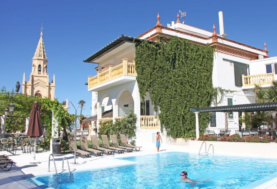 A tranquil outdoor area features a salt-water pool surrounded by lounge chairs. A building with decorative vines and balconies provides a welcoming backdrop. A historic tower, likely a church, is visible in the background under a clear blue sky.