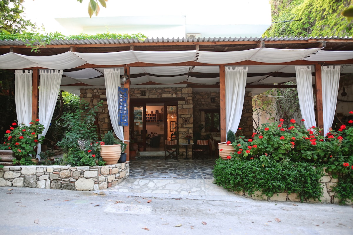 The entrance features a welcoming facade made of natural stone, accented by lush greenery and red flowers in decorative pots. Draped white fabric softens the outdoor space, creating a cozy atmosphere. Stone pathways lead to the inviting entrance of the hotel.